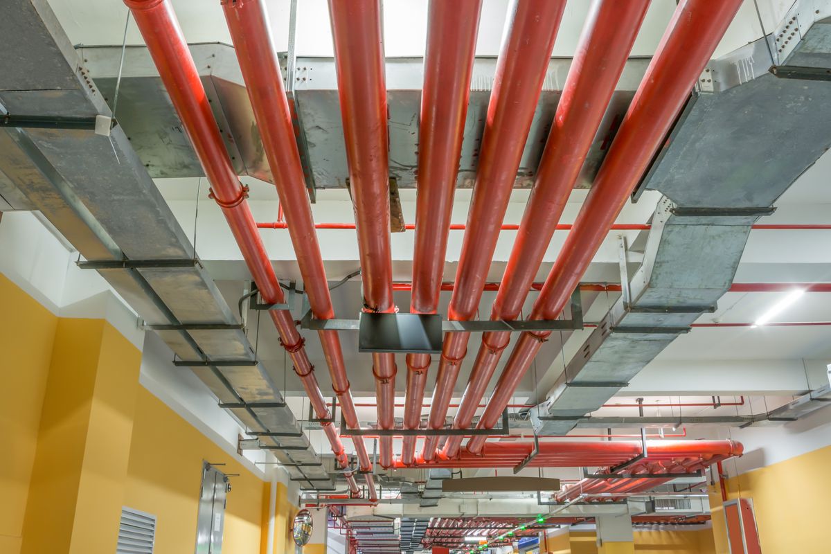 Canalisations rouges parallèles suspendues sous le plafond d'un sous-sol, entre des gaines métalliques grises.