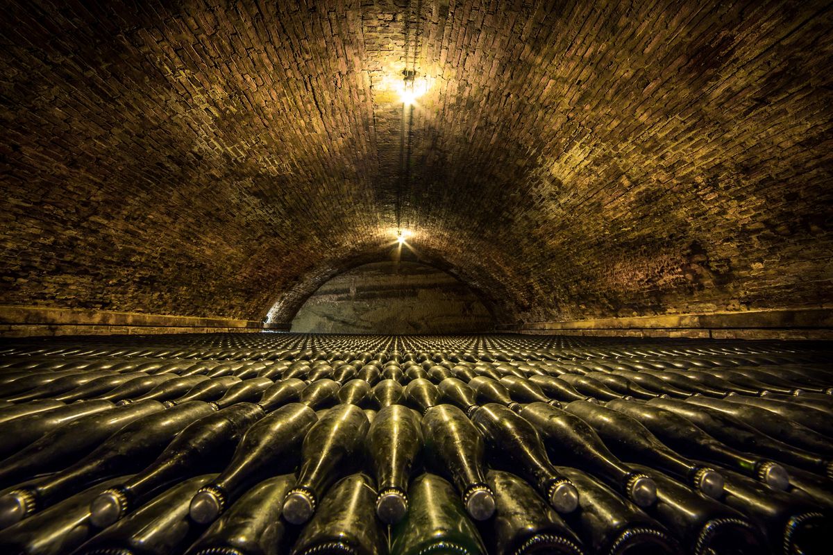 Cave voûtée en brique éclairée par deux ampoules, avec des rangées de bouteilles couchées au sol.