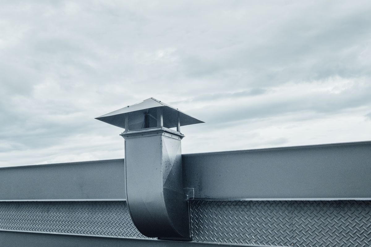Un conduit de ventilation métallique coiffé d'un chapeau dépasse d'un toit plat sous un ciel nuageux.