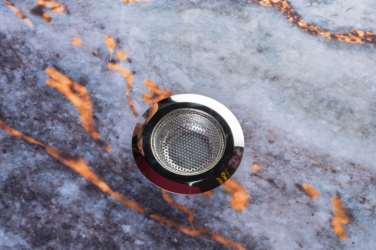 Crépine d'évier en inox à mailles fines encastrée dans un plan en marbre veiné gris et orangé.