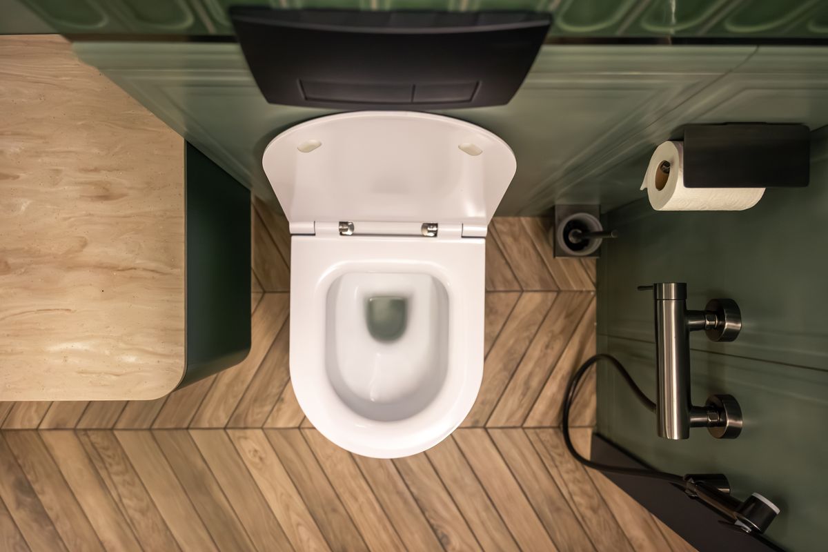 Cuvette de WC suspendue vue du dessus, abattant relevé, sur un parquet en chevrons près d'un dérouleur de papier.