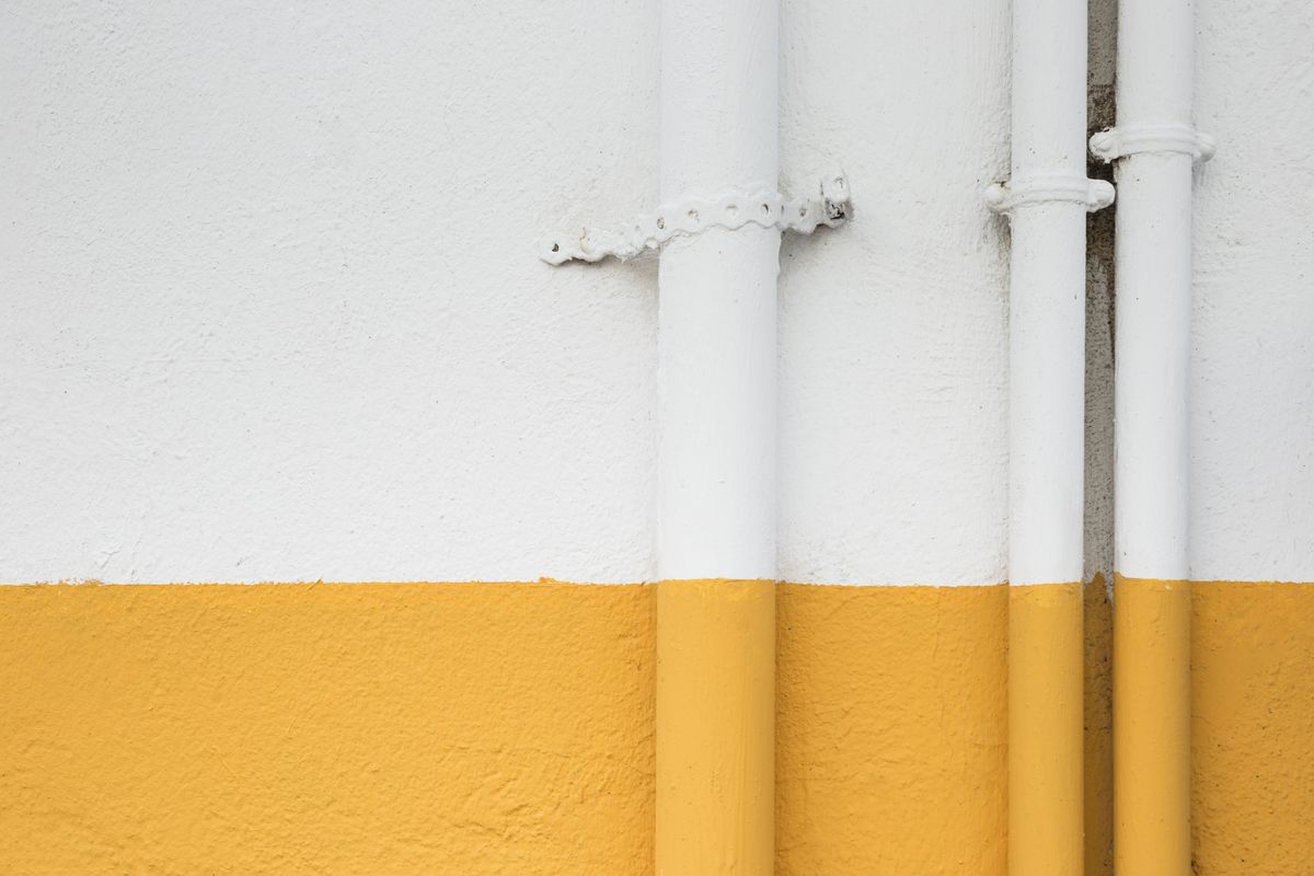 Plusieurs descentes de tuyaux blancs fixées par des colliers le long d'un mur blanc et jaune.