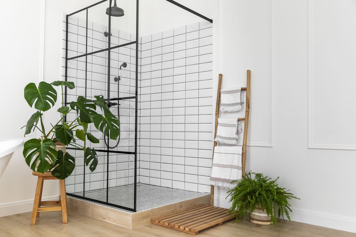 Douche à paroi vitrée cerclée de noir, carrelage blanc, plantes vertes et échelle porte-serviettes en bois.