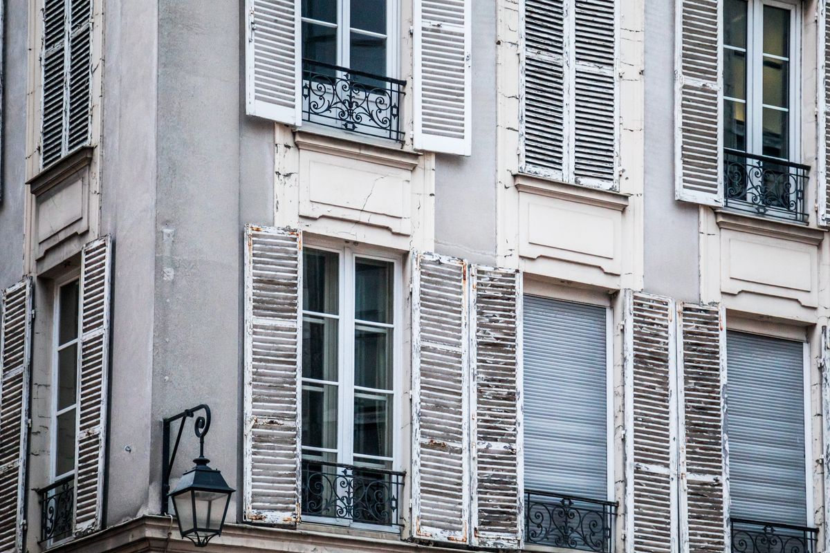 Façade d'immeuble ancien en pierre claire, avec des volets en bois ouverts et des balcons en fer forgé.