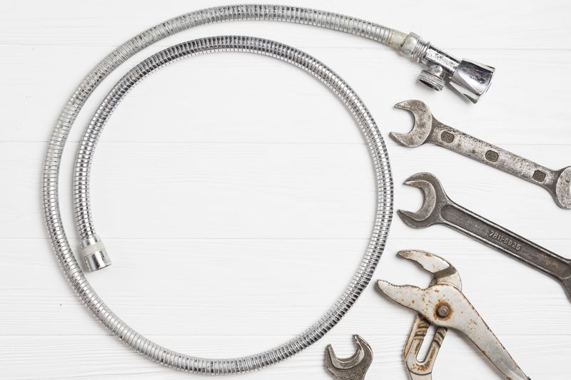 Flexible métallique enroulé, deux clés plates, une clé à molette et une pince multiprise posés sur un plan blanc