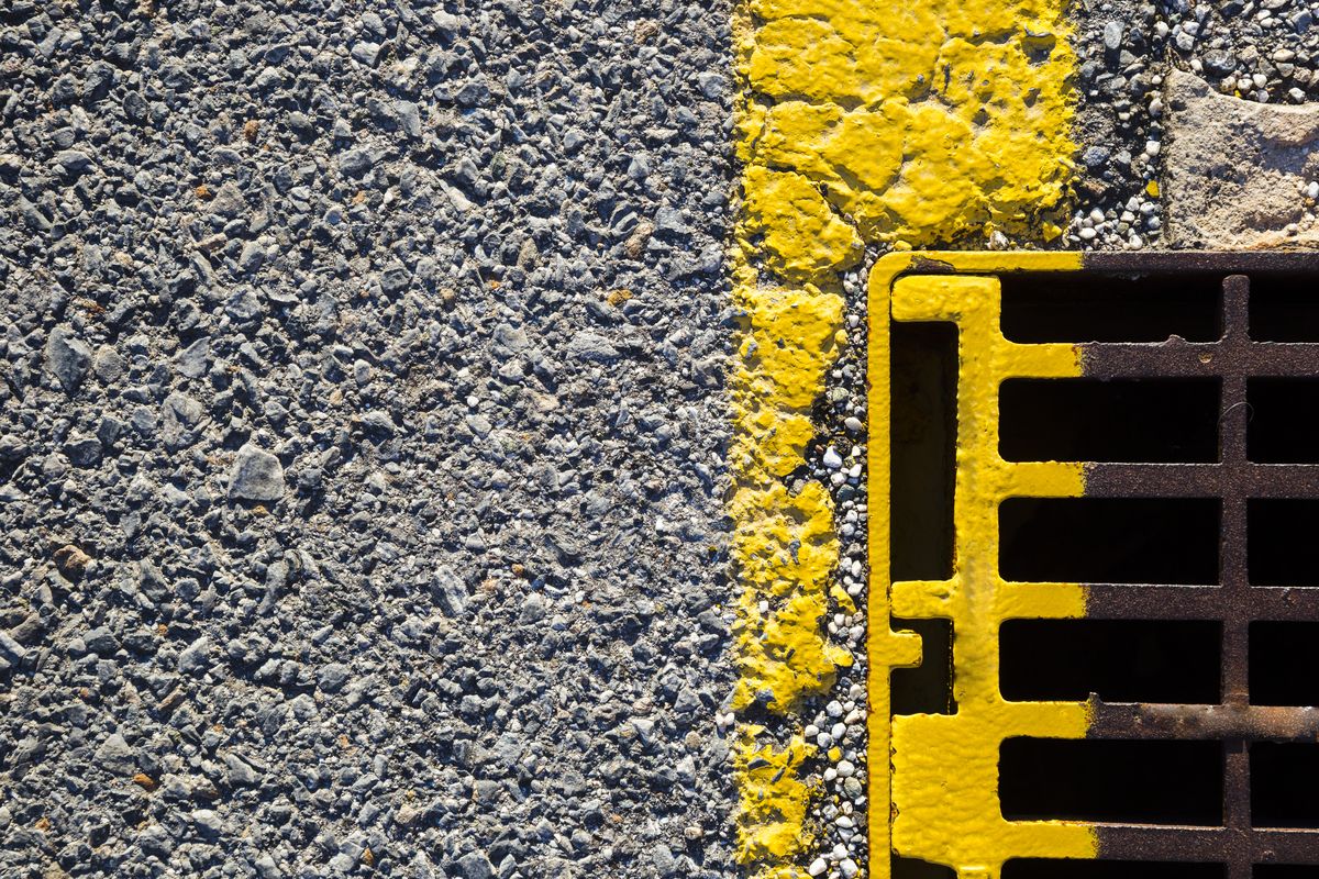 Grille d'avaloir métallique en partie peinte en jaune, au bord d'une chaussée en enrobé marquée d'une ligne jaune.