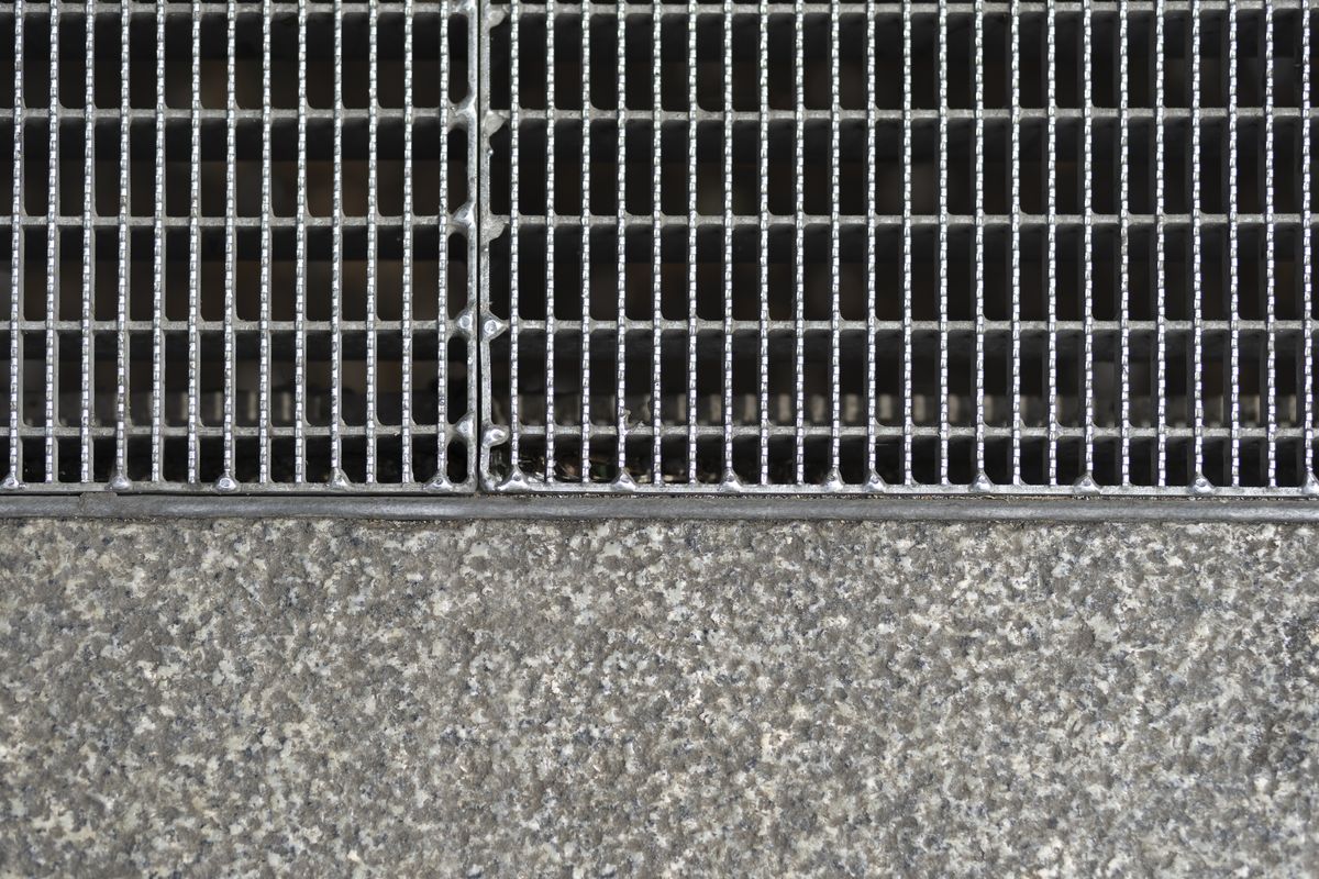 Grille métallique en caillebotis posée au ras du sol, à côté d'une dalle de béton gravillonné.