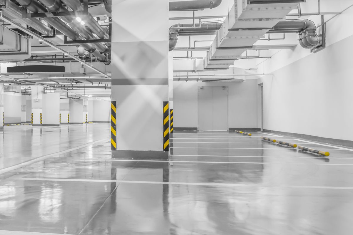 Parking souterrain vide au sol gris brillant, avec des piliers balisés de jaune et des canalisations au plafond.