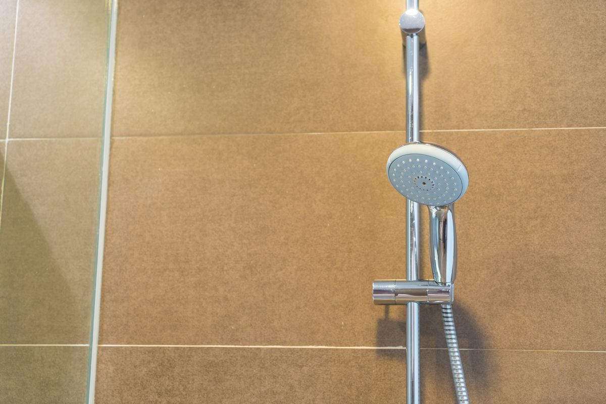 Pomme de douche chromée fixée sur sa barre, devant un mur carrelé de grands carreaux beiges.