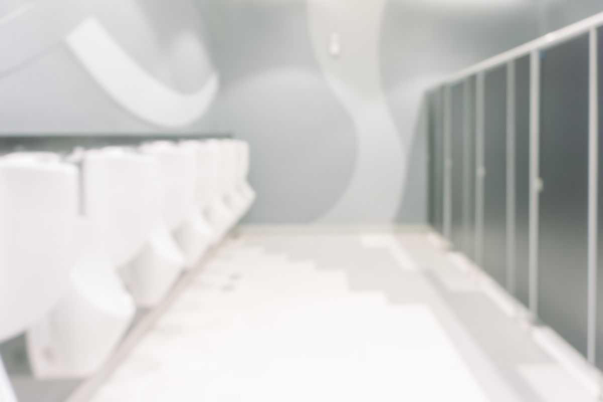 Bloc sanitaire collectif clair, avec une rangée d'urinoirs blancs face à des cabines grises, vu en flou.