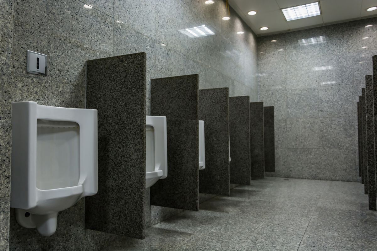 Rangée d'urinoirs blancs séparés par des cloisons sombres dans des sanitaires collectifs au sol de granit.
