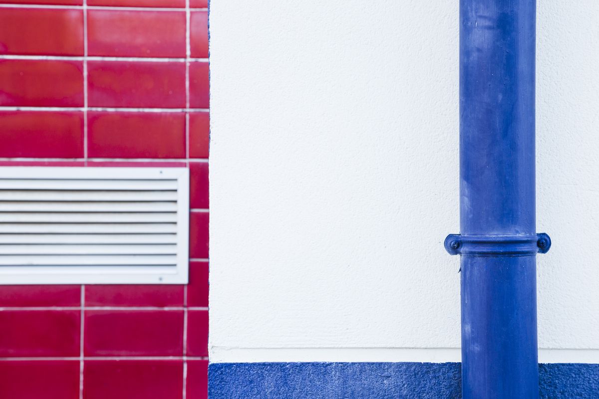 Tuyau vertical bleu fixé par un collier sur un mur blanc, à côté d'une grille d'aération.