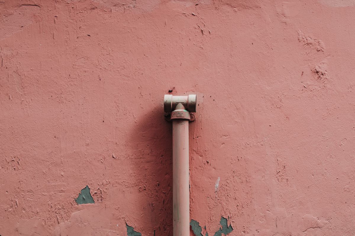 Tuyau vertical muni d'un raccord, fixé contre un mur rose dont la peinture s'écaille par endroits.