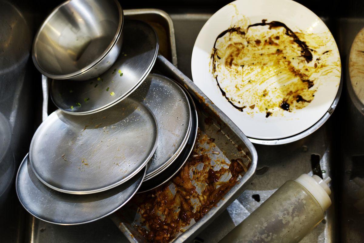 Assiettes, bols en inox et plat encore gras empilés dans le bac de plonge d'une cuisine professionnelle.