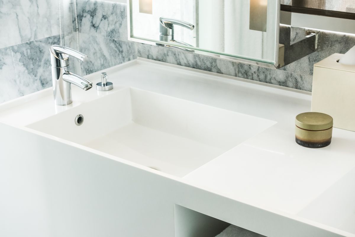 Vasque rectangulaire blanche intégrée à un plan de toilette, avec son trop-plein, un mitigeur chromé et un miroir éclairé.