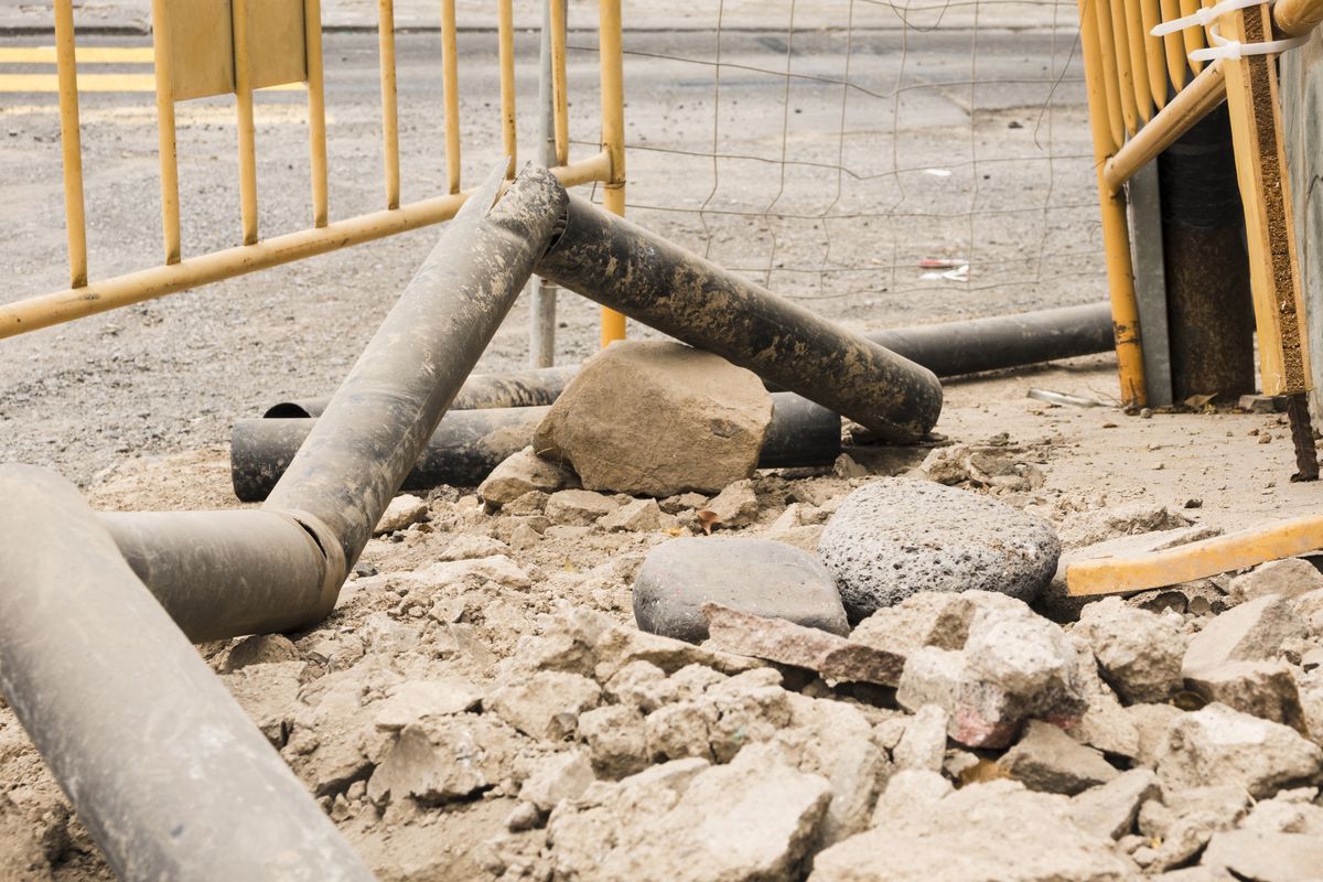 De vieux tuyaux gris gisent sur des gravats et des pierres, derrière des barrières de chantier jaunes.