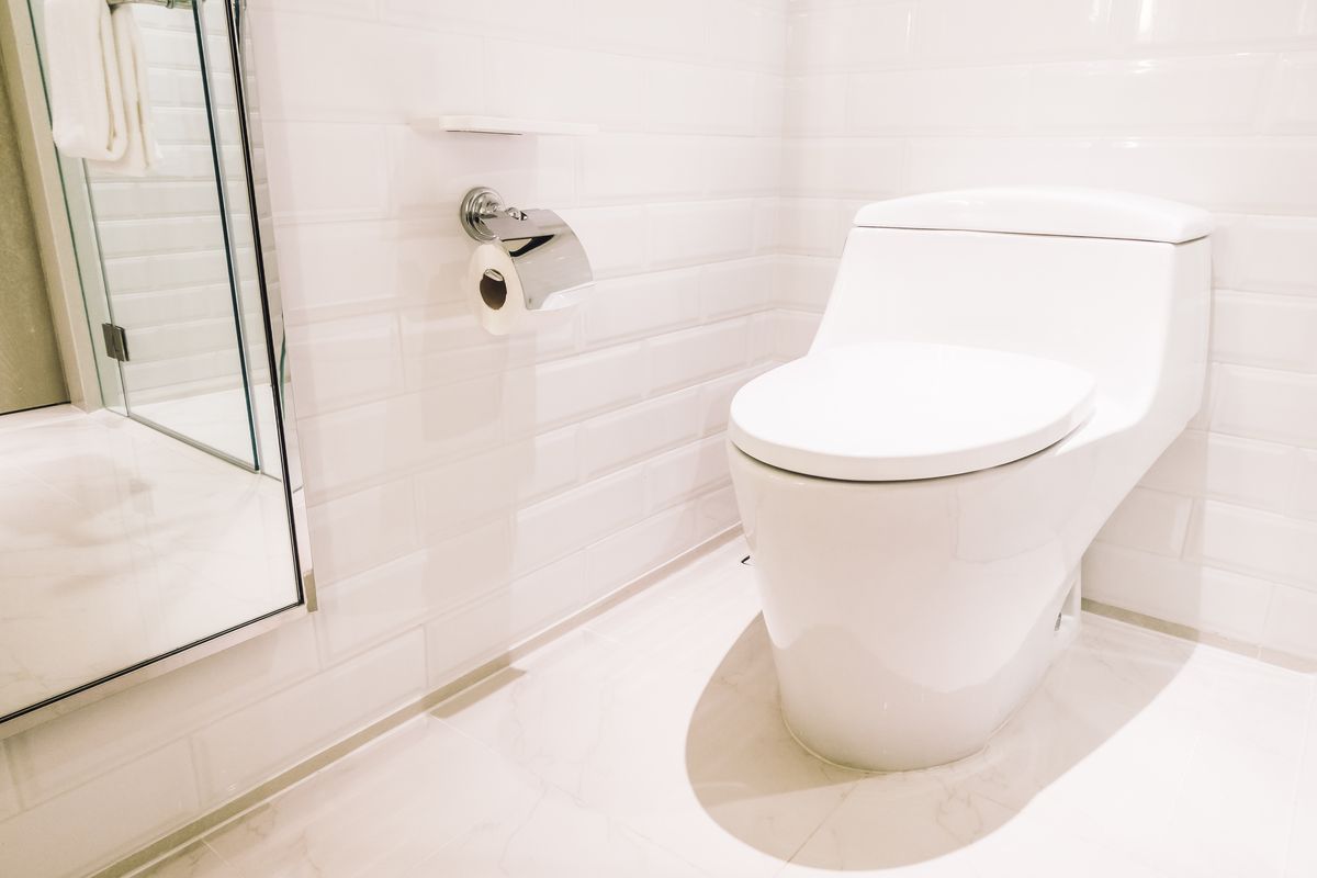 WC blanc posé au sol dans une pièce carrelée de blanc, près d'une paroi vitrée et d'un dérouleur.
